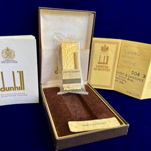 May include: A vintage Dunhill lighter in its original box, with paperwork. The gold-toned lighter is in a plastic sleeve. The box is brown with a velvet lining. Additional documents include a guarantee and a booklet.