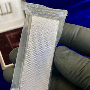 May include: A silver-colored lighter with a textured, wave-like pattern, sealed in a clear plastic bag. The lighter is rectangular and appears to be new. The background includes a brown box and a dark blue surface.