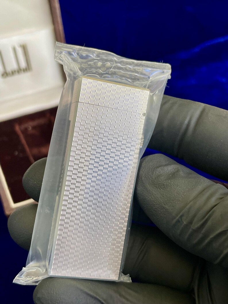 May include: A silver-colored lighter with a textured, wave-like pattern, sealed in a clear plastic bag. The lighter is rectangular and appears to be new. The background includes a brown box and a dark blue surface.