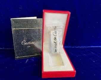 May include: A silver-colored lighter with vertical lines is displayed in a red box with a white interior. The lighter is in a clear plastic sleeve with the text "ter mut de Cartier". A black box with the Cartier logo is to the left.