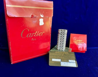 May include: A silver-toned Cartier lighter with a geometric design, accompanied by a red box and paperwork. The box features the Cartier logo in gold lettering. The background is a deep blue.
