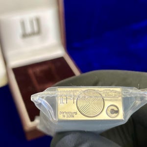 May include: A gold-colored lighter in a clear plastic wrapper. The lighter has a circular design and the text "MADE IN SWITZERLAND". The lighter is in a brown box with a white interior.