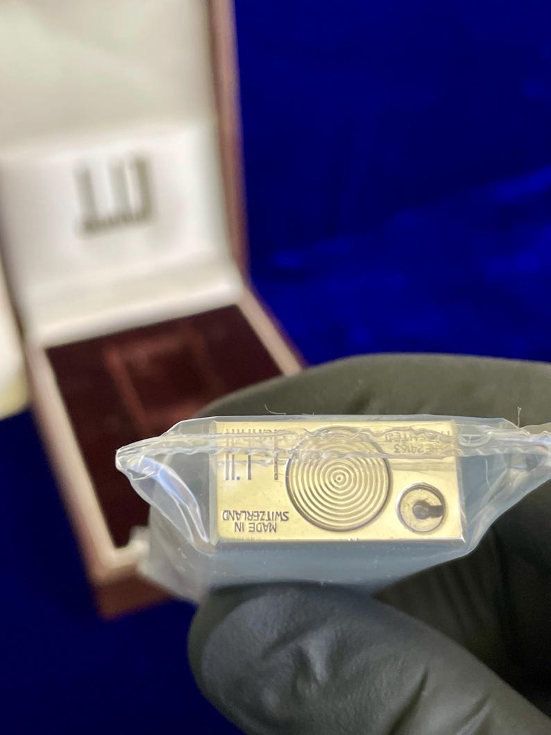May include: A gold-colored lighter in a clear plastic wrapper. The lighter has a circular design and the text "MADE IN SWITZERLAND". The lighter is in a brown box with a white interior.