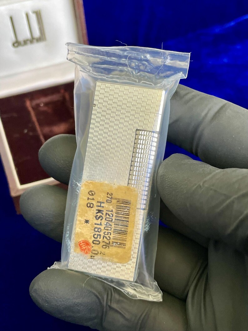May include: A silver-colored Dunhill lighter in a clear plastic bag. The lighter has a textured pattern and a rectangular shape. A price tag is visible on the lighter. The Dunhill logo is visible on the box.