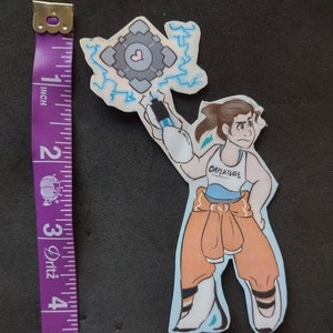 May include: A cartoon illustration of a woman wearing a blue and orange jumpsuit with the word "Aperture" on the front. She is holding a blue and white cube with a heart on it. The cube is surrounded by blue lightning bolts.