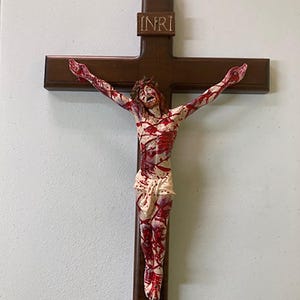 Könnte beinhalten: Ein Holzkruzifix mit einer weißen Figur von Jesus Christus, bedeckt mit roter Farbe, die Blut darstellt. Die Inschrift "INRI" befindet sich auf dem Kreuz.