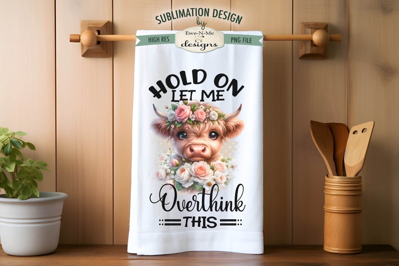 May include: A white kitchen towel featuring a brown and white Highland cow wearing a pink flower crown. The text "HOLD ON LET ME OVERTHINK THIS" is printed in black on the towel.