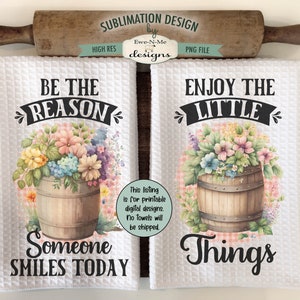 May include: Two white kitchen towels with floral designs and text. The first towel says "Be the reason someone smiles today" and features a barrel with pink, yellow, and blue flowers. The second towel says "Enjoy the little things" and features a barrel with pink, yellow, and blue flowers.