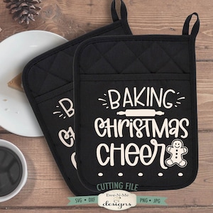 Pode incluir: Duas luvas de forno pretas acolchoadas com texto branco "BAKING CHRISTMAS CHEER" e um boneco de gengibre. As luvas têm uma argola para pendurar e um bolso. A imagem também inclui um prato com uma fatia de torta e uma xícara de café.