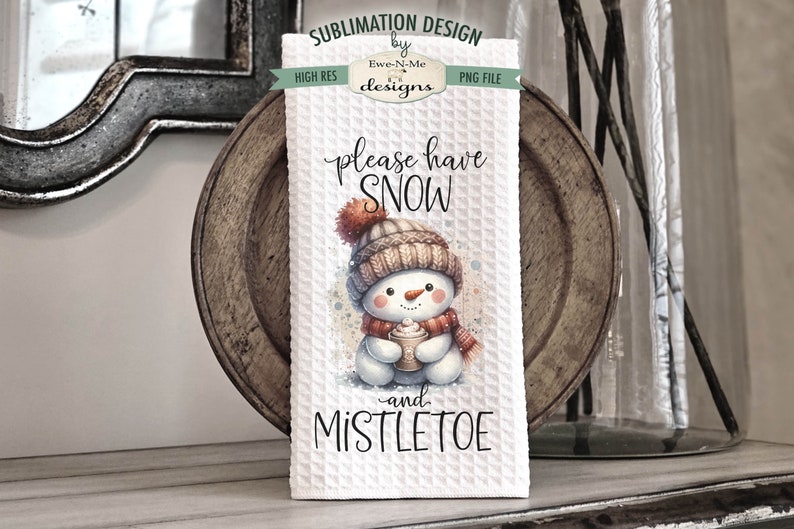 May include: White kitchen towel featuring a watercolour illustration of a snowman holding a cup of hot chocolate. The text "please have snow -and- mistletoe" is printed on the towel.