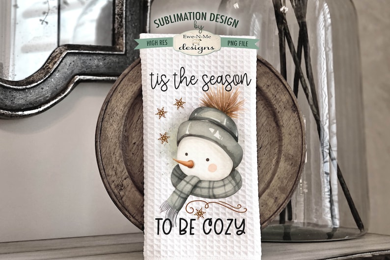 Pode incluir: Toalha de cozinha branca com um desenho de boneco de neve em aquarela. O boneco de neve est&aacute; usando um chap&eacute;u e uma echarpe cinzas. O texto "tis the season to be cozy" est&aacute; impresso na toalha.