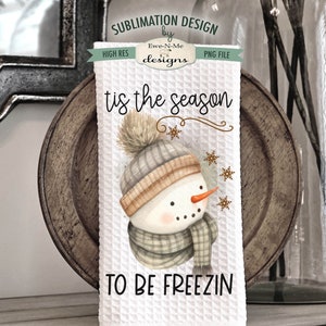 Pode incluir: Toalha de cozinha branca com uma ilustra&ccedil;&atilde;o em aquarela de um boneco de neve usando um chap&eacute;u e uma echarpe marrons e cinzas. O texto "tis the season to be freezin" est&aacute; impresso na toalha.