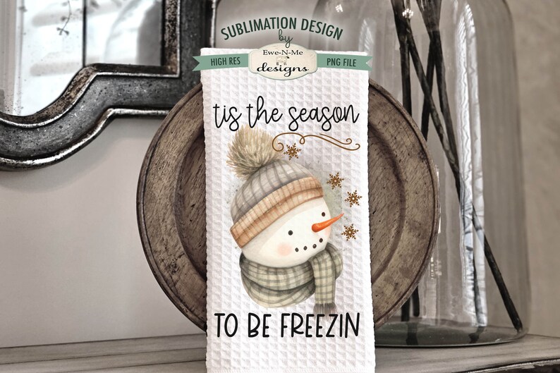 Pode incluir: Toalha de cozinha branca com uma ilustra&ccedil;&atilde;o em aquarela de um boneco de neve usando um chap&eacute;u e uma echarpe marrons e cinzas. O texto "tis the season to be freezin" est&aacute; impresso na toalha.