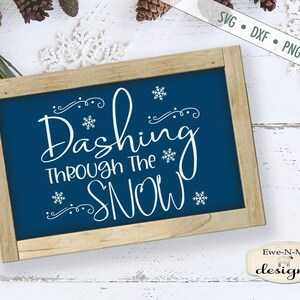 May include: A blue wooden sign with white text that reads "Dashing Through The Snow" with a decorative snowflake design. The sign is on a white wooden background.