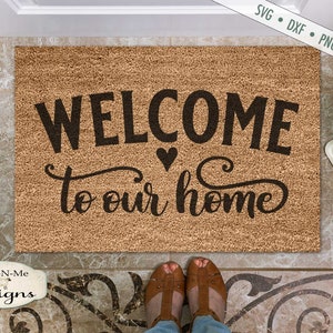 以下が含まれることがあります： 天然ココナッツ繊維で作られた長方形のドアマット。「WELCOME to our home」という言葉が黒色の装飾的なフォントで、ハートのシンボルとともに表示されています。デザインには「SVG • DXF • PNG • JPG」というテキストが含まれています。Ewe-N-Me designsのロゴが見えます。