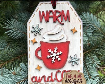 Op de afbeelding: Houten lasergesneden tag-ornament met de tekst "WARM and COZY". De tag heeft een rode mok met slagroom, sneeuwvlokken en sterren. Het ornament is ontworpen met een rustieke esthetiek en bevat de tekst "EWE-N-ME designs".