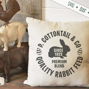 Puede incluir: Una almohada blanca con un dise&ntilde;o circular negro que dice "P. Cottontail & Co. Quality Rabbit Feed Since 1923 Premium Blend". El dise&ntilde;o tambi&eacute;n incluye una silueta de un conejo y dos huevos de Pascua.