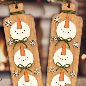 May include: Two wooden cutting boards with a snowman design. Each board features three white snowman faces with orange carrot noses and black dot eyes. Green twine bows and snowflake accents add detail. The text "LASER-READY SNOWMAN CUTTING BOARD DESIGN" is at the top.