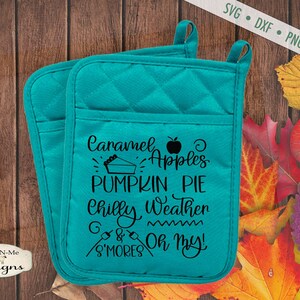 Puede incluir: Dos agarraderas acolchadas turquesas con el texto "Caramel Apples, Pumpkin Pie, Chilly Weather & S'mores Oh My!" en escritura negra. Las agarraderas tienen un lazo para colgar y un bolsillo. El fondo incluye hojas de otoño.