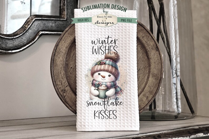 May include: A white kitchen towel with a winter-themed design featuring a snowman holding a cup of hot chocolate. The text "winter wishes snowflake kisses" is printed on the towel.