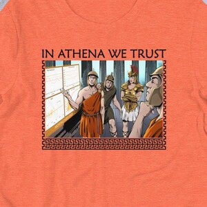 以下が含まれることがあります： オレンジ色のTシャツに、古代ギリシャの服装をした人々のグラフィックがプリントされています。Tシャツには「IN ATHENA WE TRUST」という文字がプリントされています。