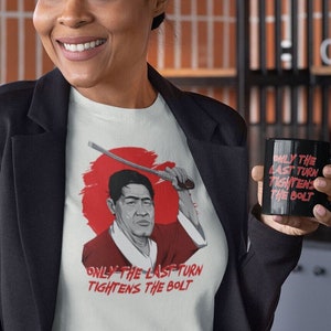 May include: A white t-shirt with a red and black illustration of a man holding a sword. The text "Only the last turn tightens the bolt" is printed on the shirt. The person in the image is holding a black mug with the same text printed on it.