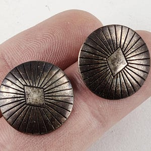 May include: Two antique silver-toned metal buttons. Each button is round with a central square design and radiating lines. The buttons are held between fingers, showcasing their size and detail.