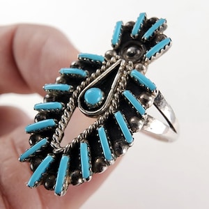 May include: A silver ring with a large, ornate design. The ring features turquoise-colored rectangular stones and a central teardrop shape with a turquoise stone. The ring has a detailed, textured border and a classic band.