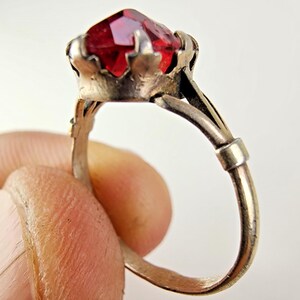 May include: A silver-toned metal ring featuring a faceted, deep red gemstone set in a crown-like prong setting. The band is thin and includes a small, rectangular metal detail on the side, showcasing a metallic finish with visible signs of wear.