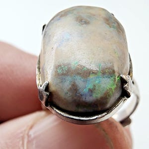 May include: A silver ring featuring a large, oval-shaped opal gemstone. The opal displays a mix of colors, including green, blue, and brown, set in a silver band. The ring is held up by a finger.