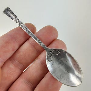 May include: A small silver-toned metal spoon featuring an elongated handle with intricate geometric engravings, including diamond and cross-hatch patterns. The bowl of the spoon displays a smooth, reflective surface with a decorative floral motif at the base.