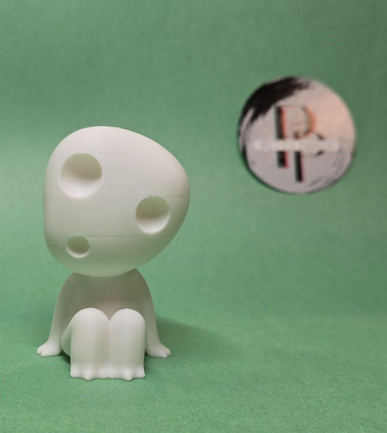 May include: White 3D printed figurine of a character with a large head and three holes. The character is sitting with its legs crossed.