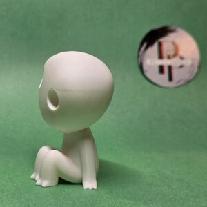 May include: A white, 3D printed figurine of a small, round-headed creature with two holes in its head. The creature is sitting with its legs crossed.