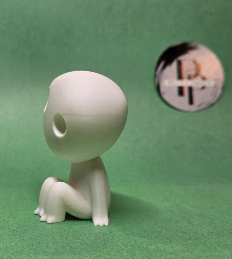 May include: A white, 3D printed figurine of a small, round-headed creature with two holes in its head. The creature is sitting with its legs crossed.