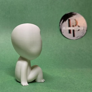 May include: A white, 3D printed figurine of a person sitting with their legs crossed. The figurine has a large, round head and no facial features.