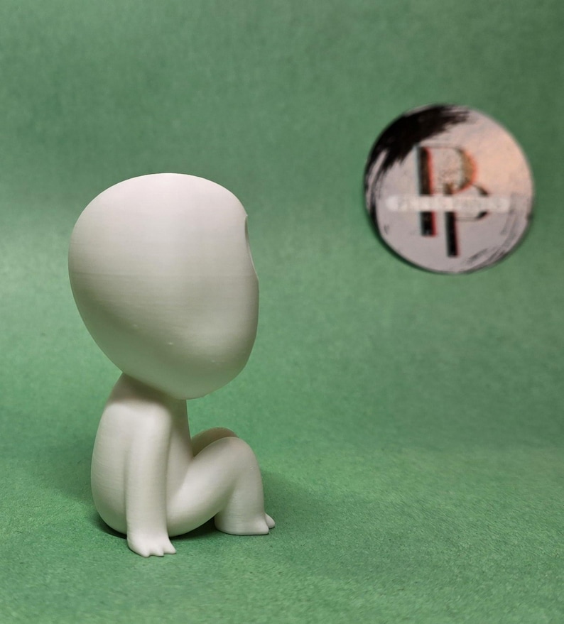 May include: A white, 3D printed figurine of a person sitting with their legs crossed. The figurine has a large, round head and no facial features.
