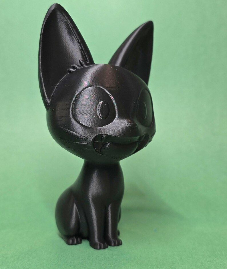 May include: A black 3D printed figurine of a cartoon cat with large eyes and a wide smile. The cat is sitting with its legs tucked under its body.