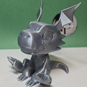 May include: A 3D printed gray figurine of a cartoon dragon with large ears and wings. The dragon has a friendly expression and is sitting with its legs crossed.