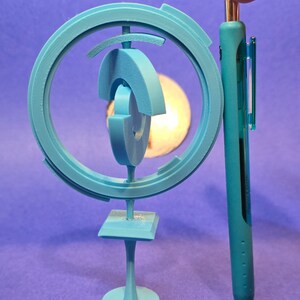 May include: A light blue 3D printed sculpture with a circular design and a base. The sculpture is about the size of a pen.