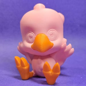 May include: A pink 3D printed bird figurine with orange beak and feet. The bird has a cute, cartoon-like appearance.