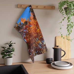 Peut inclure: Un torchon de cuisine rectangulaire suspendu à un porte-ustensiles en bois, arborant une impression photographique vibrante d'arbres en automne avec un feuillage orange, rouge et jaune sur un ciel bleu vif, dans une cuisine avec des plantes.