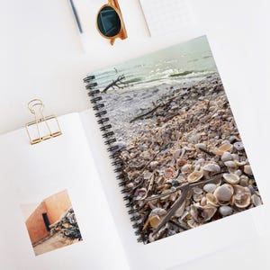 Puede incluir: Cuaderno de espiral con una escena de playa, con conchas marinas y madera flotante. La imagen muestra un primer plano de las conchas y el océano. El cuaderno está abierto sobre una superficie blanca, con un clip dorado y gafas de sol.