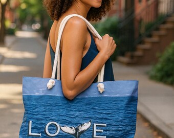 Könnte beinhalten: Blaue Tragetasche mit weißen Seilgriffen. Die Tasche zeigt das Wort "LOVE" mit einem Walflossendesign. Der Hintergrund zeigt eine Meereslandschaft. Die Tasche wird von einer Person getragen, die einen Strohhut und ein blaues Oberteil trägt.