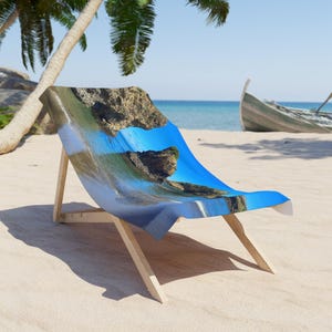 May include: A beach chair with a vibrant blue and white beach scene printed on the fabric. The chair has a natural wood frame and is set on a sandy beach with a palm tree and a boat in the background.