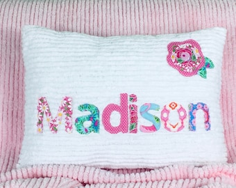 May include: A white rectangular pillow with a textured ribbed surface featuring the name Madison appliqued in colorful floral and polka dot fabric letters. A matching floral fabric flower applique is positioned in the top right corner.