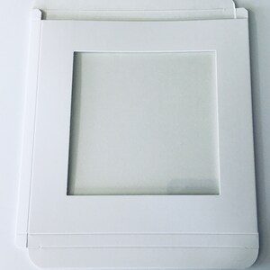 May include: White cardboard box with a square window cut out. The box is designed to hold a square item, such as a photo or a piece of art.