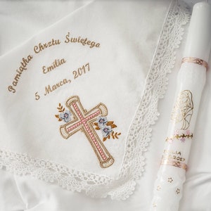 May include: A white handkerchief with embroidered cross and floral details, and text in gold thread. A white candle with gold accents and a religious illustration is also present. The text on the handkerchief reads "Pamiatka Chrztu Swietego Emilia 5 Marca, 2017".
