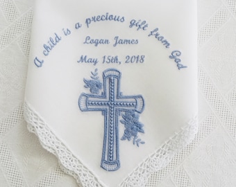 Puede incluir: Un pañuelo blanco bordado con una cruz azul y un diseño floral. El texto dice "A child is a precious gift from God" con el nombre "Logan James" y la fecha "May 15th, 2018".