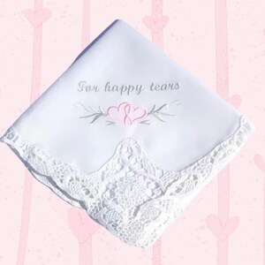 May include: White handkerchief with embroidered text "For happy tears" and a floral design with two pink hearts. The handkerchief has a delicate lace trim along the edge. The background is a pink pattern with hearts.