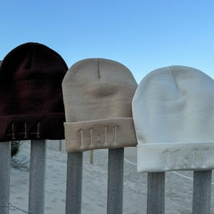 May include: Three beanies in shades of brown, tan, and cream are displayed on a wooden fence. The brown beanie has the number "444" embroidered on the front, while the tan and cream beanies have "11:11" embroidered on the front. The background shows a beach and blue sky.